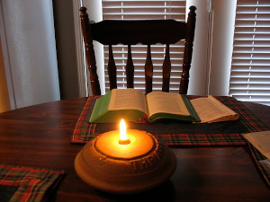 An empty chair at dining room table with a lit yellow candle in the center, and open book of Scripture in front of the chair with a lit window closed blinds in the background.