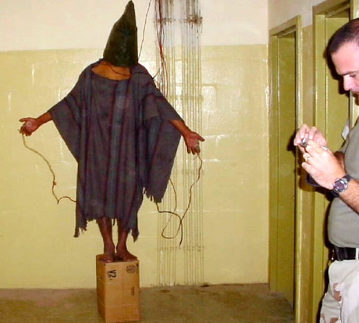 A barefoot and hooded prisoner at Abu Ghraib standing on a box with wires attached to the fingers on both hands with an US Military man in the foreground.