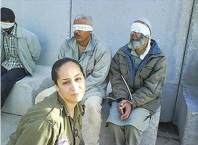 Smiling female Israeli soldier in the foreground with thee bound and blindfolded prisoners sitting on stone blocks in the background.