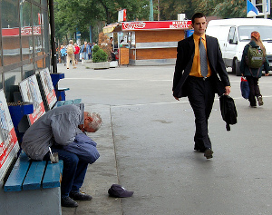 A well dressed middle aged man wearing a suit and tie and carrying a briefcase, walking past a white haired man, sitting on a bench on a city street.