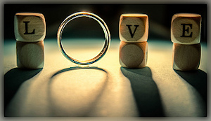 3 blocks of wood with the letters L, V, & E with a ring between the letters L and VE on a dark surface with a light source behind them.