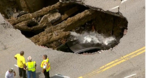 Construcion workers gathered arounda huge sinkhole on a four lane road.